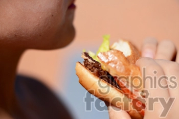 The photo shows a person eating a hamburger, which is a sandwich made of a grilled patty of ground beef served on a bun with various toppings like lettuce, tomato, cheese, and condiments such as ketchup and mustard. The person appears to be holding the hamburger with both hands, taking a bite out of it, and enjoying their meal.
